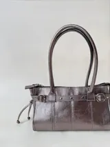 Cartera de cuero de tamaño medio con estructura rígida. Presenta una tira decorativa con tachas y hebillas metálicas en los extremos, además de lazos laterales. Cuenta con doble asa para llevar al hombro y cierre superior.