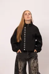 Campera negra de cuello alto con cierre frontal de cremallera y solapa con botones metálicos. Presenta mangas largas con puños elásticos y diseño vanguardista.