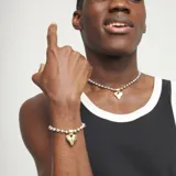 Pulsera de eslabones tipo bola de aleación de metales bañada en plata, con un dije central en forma de corazón tridimensional bañado en oro 18k. Incluye un cierre de mosquetón y una pequeña etiqueta grabada con el logo de la marca.