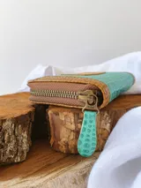 Billetera compacta de cuero con diseño bicolor en turquesa y marrón, con textura tipo reptil. Cuenta con cierre metálico perimetral y parche frontal con el logo de la marca.