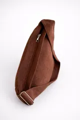 Bolso tipo bandolera de tamaño mediano, confeccionado en material con acabado efecto gamuza color chocolate. Presenta un diseño minimalista con cierre superior de cremallera y correa larga regulable con herrajes metálicos.