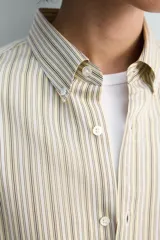 Camisa de algodón de corte relajado, con cuello solapa abotonado (button-down) y manga larga terminada en puño con botón. Presenta un patrón de rayas verticales finas en tonos beige claro y marrón/verde oliva.