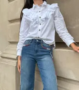 Camisa blanca de algodón con cuello clásico, cierre frontal de botones y detalle de volados en la parte superior delantera y hombros.