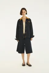 Chaqueta oversize de denim azul oscuro con cuello camisero de pana color mostaza, cierre frontal con botones metálicos y bolsillos delanteros con solapa. Cuenta con detalle de bordado en el pecho.