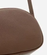Cartera tipo camera bag pequeña, color marrón camel, de forma ovalada con superficie texturizada. Cuenta con correa ajustable larga, cierre principal con cremallera dorada y un pequeño bolsillo frontal con cremallera.
