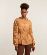 Campera deportiva femenina tipo parka, color ocre, confeccionada en poliamida y poliéster. Tiene cuello con capucha, mangas largas con puños elásticos, cierre frontal con cremallera y cordón ajustable en la cintura.