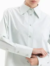 Camisa de poplin color crema, de corte clásico con cuello camisero, mangas largas y cierre frontal con botones dorados de media esfera.