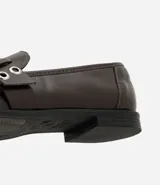 Mocasín femenino de cuero sintético color marrón oscuro, con punta redondeada y suela plana. Presenta un diseño con tiras cruzadas en el empeine decoradas con ojales metálicos y una hebilla ajustable.