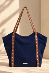 Cartera tipo tote de gamuza color azul marino, con asas dobles de cuero marrón texturado y detalles de tachas metálicas plateadas. Cuenta con una placa metálica frontal con el logo de la marca Pecarí.