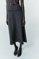 Vestido midi de tejido texturizado en color gris oscuro, con cuello alto fluido, diseño sin mangas y acabados deshilachados. Incluye un cinturón fino de cuero en color marrón con hebilla metálica y cierre de cremallera oculta en la espalda.