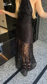 Vestido largo de encaje negro con cuello halter y espalda descubierta. Tiene un corte a la altura de la rodilla y un ruedo largo transparente.