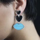 Par de aros colgantes asimétricos con forma de corazón y labios. Uno de los aros tiene un corazón grande plateado y un labio celeste con borde plateado. El otro aro tiene un labio celeste con borde plateado y un corazón pequeño plateado.