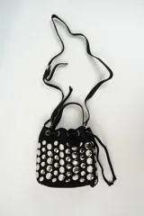 Cartera tipo bucket bag de gamuza negra, decorada con múltiples tachas metálicas plateadas de forma circular. Cuenta con cierre de cordón ajustable y asa superior.