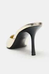 Sandalia mule de tacón alto fino y acabado en punta, con cuerpo de efecto metalizado en color dorado y tacón en color negro.