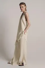 Top halter color crudo, de escote en V y diseño confeccionado en hilatura con mezcla de viscosa. Presenta bajo asimétrico con flecos al tono y cierre en la espalda.