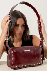 Cartera de hombro color bordó, confeccionada en cuero con acabado brillante. Presenta detalles de remaches metálicos plateados en los laterales y la base, y una correa ajustable con hebilla. Incluye una placa metálica con el logo de la marca en la parte frontal.