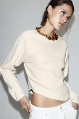 Jersey de punto negro de manga larga con cuello redondo decorado con aplicación de bolas doradas. Presenta un diseño de bajo asimétrico y cierre posterior con abertura y botón.