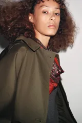 Trench corto color verde militar, con cuello solapa y capucha. Tiene manga larga con trabillas y botón en los puños, bolsillos delanteros y bajo asimétrico. Cierre frontal con botones. El tejido es repelente al agua.