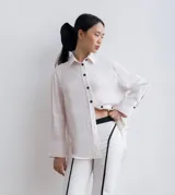 Camisa de lino color blanco roto, de corte holgado y manga larga con puños anchos. Presenta botones negros en el frente y aberturas laterales.