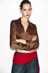 Camisa corta de tejido satinado color marrón, con cuello solapa, escote en pico y manga larga. Presenta cierre frontal con botones forrados a tono.
