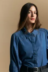 Camisa de denim azul con cuello camisero, cierre frontal de botones y mangas largas abullonadas con hombros fruncidos.