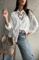 Camisa blanca de corte clásico con cuello camisero y cierre frontal de botones. Presenta un delicado bordado tono sobre tono en las mangas y una textura sutil en la tela.