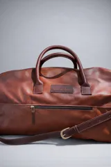 Bolso de viaje tipo weekender de cuero color tostado con asas y detalles en marrón oscuro, herrajes metálicos y bolsillo frontal con cierre.