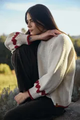 Sweater de punto color crudo con diseño de corazones rojos en las mangas, cuello redondo y terminaciones acanaladas en contraste rojo.