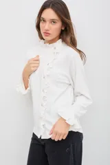 Remera de manga larga color blanco, confeccionada en tejido liviano con caída fluida. Presenta cuello alto con volados, abotonadura frontal decorativa y detalles de volados a lo largo de la abertura, además de terminaciones con volados en los puños.