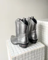 Bota estilo tejana de cuero vacuno en color plateado metalizado, con taco bajo cuadrado y costuras decorativas en la puntera.