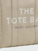 Bolso tipo tote de gran tamaño en color beige, confeccionado en lona resistente. Presenta el logo distintivo de la marca estampado en la parte frontal con letras blancas. Cuenta con doble asa superior para llevar al hombro y cierre superior con cremallera.