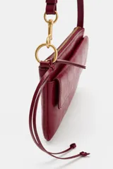 Bolso de hombro color bordó, con asa de hombro, bolsillo exterior con solapa y cierre de imán, y cierre principal con cremallera. Incluye detalle de anilla metálica dorada y tira con nudos en el lateral.