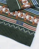 Buzo tejido de punto con diseño de patrones geométricos y rombos en tonos verde, naranja, blanco y azul. Presenta cuello redondo, puños y ruedo acanalados, con etiqueta de marca en el pecho.