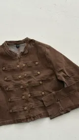 Chaqueta de estilo militar en color marrón, con corte estructurado y cuello mao. Presenta cierre frontal con cremallera y detalles de filas horizontales con botones metálicos decorativos en el frente y en los puños.