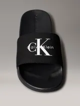 Ojotas tipo slide Calvin Klein Jeans, color negro, con banda superior de lona que presenta el logo de la marca bordado en blanco. Posee suela de EVA con plantilla ergonómica moldeada y el nombre de la marca grabado en el lateral.