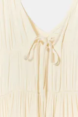 Vestido largo satinado color crudo, con escote en pico adornado con lazada y tirantes finos ajustables. Presenta detalles de frunces y diseño fluido.