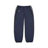Pantalón deportivo Supreme de corte holgado, color azul marino, con franjas laterales negras que incluyen el logo de la marca repetido. Presenta cintura elástica y puños ajustados en los tobillos.