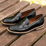 Mocasín de cuero vacuno negro con textura grabada tipo reptil. Presenta un diseño clásico con antifaz en el empeine, suela de goma en tono natural y taco bajo de madera.