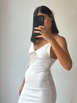 Vestido largo blanco, ajustado al cuerpo, con escote pronunciado en V y tirantes finos. Presenta un detalle de encaje o puntilla en el lateral del busto.