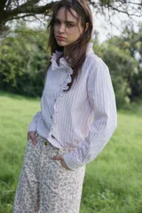 Camisa de algodón con estampado de rayas verticales en tonos beige y blanco. Presenta un diseño de manga larga, cuello camisero y un detalle de volado frontal a lo largo de la botonadura.