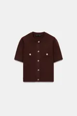 Chaqueta de punto color marrón oscuro, con cuello redondo y manga corta. Presenta solapas decorativas con botón dorado en el pecho y cierre frontal con botones dorados.