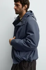 Parka de tejido técnico con acabado engomado, cuello subido y capucha ajustable. Presenta cierre frontal con cremallera oculta bajo solapa con botones a presión, bolsillos laterales de vivo y mangas largas con puños ajustables.