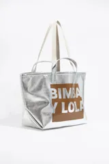 Bolso tipo tote de gran tamaño en color plateado metalizado, con acabado texturizado. Presenta un diseño rectangular con un panel central cuadrado en color marrón que incluye el logo de la marca estampado en letras plateadas. Cuenta con doble asa de mano y correas largas adicionales para llevar al hombro.