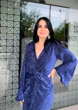 Vestido midi de fiesta en color azul, confeccionado en tela con textura satinada. Presenta un diseño cruzado con escote en V, mangas largas con puños y un detalle de fruncido en la cintura que crea una abertura frontal en la falda.