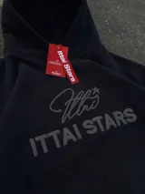 Hoodie de color negro con diseño de estampa frontal que combina una tipografía manuscrita con el texto ITTAI STARS en letras mayúsculas. Presenta un corte holgado, capucha amplia y bolsillo tipo canguro.