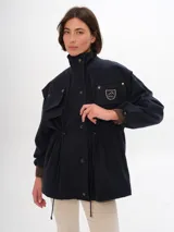 Trench azul marino con cuello alto, cierre frontal de botones a presión y cordón ajustable en la cintura. Presenta cuatro bolsillos frontales con solapa y un parche con el logo de la marca en el pecho.