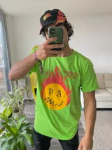 Remera verde con estampa de un círculo amarillo con llamas rojas y la inscripción "PA" en la parte superior y una sonrisa en la parte inferior.