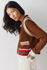 Chaleco de lana color marrón con cuello estilo bebé, cierre frontal con botones grandes y detalle de puntilla en cuello, sisas y bolsillo frontal. Presenta una franja decorativa en color rojo en el ruedo.