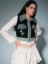 Chaleco de punto negro con cierre frontal, cuello y bordes en tejido jaspeado gris y blanco. Presenta un diseño bordado de flores coloridas en el frente.