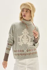 Polera de tejido de punto en color gris, con cuello alto y diseño holgado. Presenta un aplique central en color crema con motivos ornamentales y detalles bordados geométricos en la parte inferior y en las mangas.